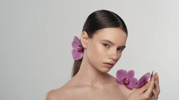 Portrait of brunette female model. Close up shot of a woman with ponytail holding an orchid flower in her hand and looking away from the camera. Organic skincare advertisement concept.