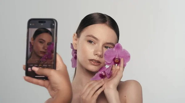 一个漂亮的模特在智能手机上摆出一副自然妆容 拿着一朵兰花 开心地笑着拍照的照片 社交媒体广告概念 — 图库照片