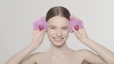 Esmer kadın modelin portresi. Bir kadının gözlerini orkide çiçekleriyle kapayarak gülümseyerek mutlu ve göz kırparken çekimini yakından çek. Organik cilt tanıtım konsepti. Ağır çekim. HDR