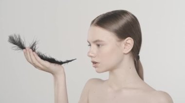 Güzel bir kadının portresi. Siyah tüy tutan bir kadın modelin yakın çekimi. Avucundaki tüyü üflüyor. Beyaz arka planda izole edilmiş. Cilt bakımı reklam konsepti. Yavaş çekim