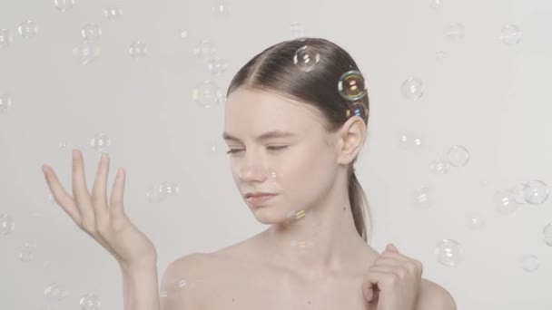 近照一个年轻女性的自然妆容 微笑着看着身边飞舞的肥皂泡 被白色背景隔离 瘦小的美容美发理念 慢动作 — 图库视频影像