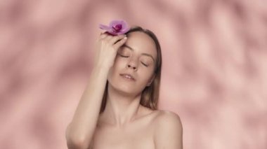 Bir kadın orkide çiçeğine dokunup temiz, pürüzsüz yüzüne dokunur. Stüdyoda elinde orkide çiçeğiyle pembe arka planda gülümseyen bir kadının portresi. Güzellik kavramı
