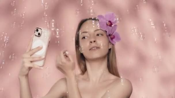 一个女人 光着肩膀 满头兰花 带着一个自拍 富丽堂皇的女人手里拿着智能手机 四周都是粉红背景的肥皂泡 — 图库视频影像