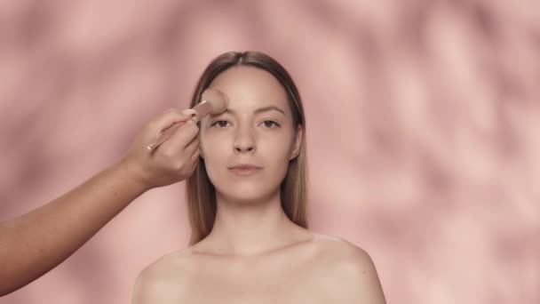 メイクプロセス メイクアップアーティストの手は ブラシで顔に女性の化粧を適用します ピンクの背景にあるスタジオのセミノードの女性 粉末を塗布する ブラシをかけます フェイシャル化粧品 コンセプト — ストック動画