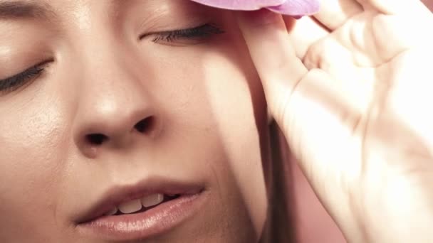 一个年轻女人的脸 皮肤洁白 背景是粉色的 一个女人把兰花摸到她的脸上 护肤的概念 慢动作 — 图库视频影像
