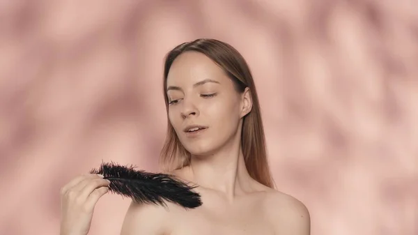 女性は黒い羽を持つ彼女の裸の肩に触れます 若い女性はピンクの背景にスタジオで滑らかで柔らかい肌を示しています ケアのコンセプト — ストック写真
