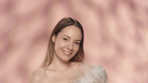 女性は白い羽を持つ彼女の裸の肩に触れます 若い女性はピンクの背景にスタジオで滑らかで柔らかい肌を示しています ケアのコンセプト — ストック写真