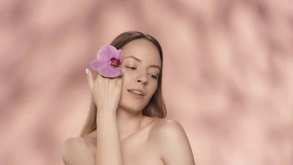 女性は彼女の顔のきれいな肌に蘭の花に触れます ピンクのバックグラウンドでスタジオでオーキッドフラワーを手に入れた女性の肖像画 美のコンセプト 化粧品 — ストック写真