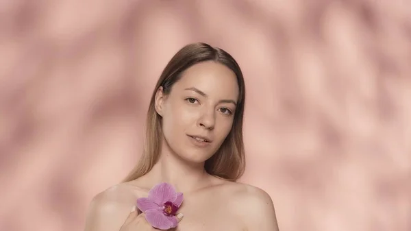 女性はきれいで滑らかな肌に蘭の花に触れます ピンクのバックグラウンドでスタジオでオーキッドフラワーを手に入れた女性の肖像画 化粧品 肌のコンセプト — ストック写真