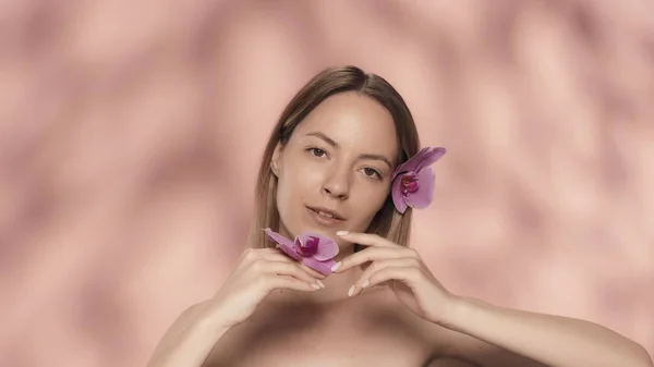 女性は蘭の花のベルベットの花びらに触れます 彼女の髪に蘭の花を咲かせた女性と ピンクの背景に手を差し伸べた女性 スキンケアのコンセプト — ストック写真