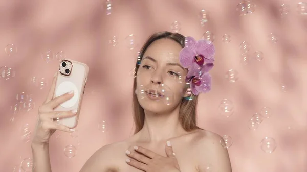 一个女人 光着肩膀 满头兰花 带着一个自拍 富丽堂皇的女人手里拿着智能手机 四周都是粉红背景的肥皂泡 — 图库照片