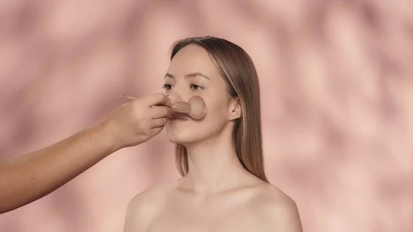 メイクプロセス メイクアップアーティストの手は ブラシで顔に女性の化粧を適用します ピンクの背景にあるスタジオのセミノードの女性 粉末を塗布する ブラシをかけます フェイシャル化粧品 コンセプト — ストック写真