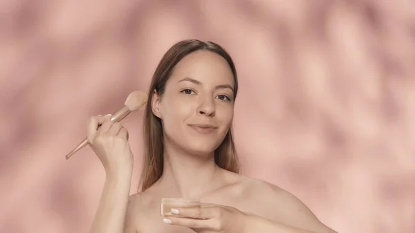 ピンクのバックグラウンドでスタジオに化粧ブラシと粉の瓶を持つ女性 粉末を塗布する ブラシをかけます スキンケアのコンセプト — ストック写真