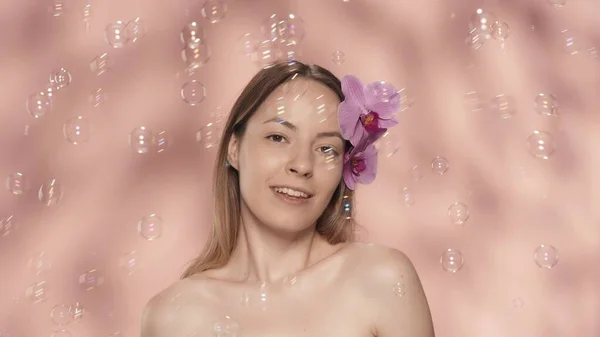 女性らしさ 彼女の髪に裸の肩と蘭の花を持つ美しい官能的な女性の肖像画 ピンクの背景に石鹸の泡に囲まれたスタジオの女性 美のコンセプト — ストック写真