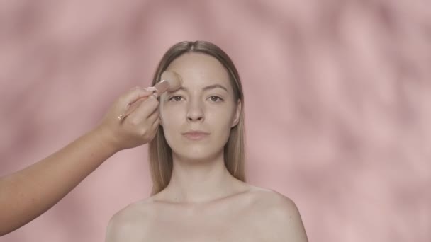 メイクプロセス メイクアップアーティストの手は ブラシで顔に女性の化粧を適用します ピンクの背景にあるスタジオのセミノードの女性 粉末を塗布する ブラシをかけます 美のコンセプト — ストック動画