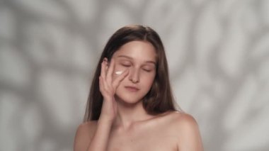 Çekici beyaz bir modelin portresi. Bir kızın kameraya bakıp yüzüne ve yanaklarına nemlendirici krem sürerken yakın çekim. Güzellik derisi reklam konsepti.