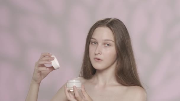 一个持有化妆品的年轻模特的画像 女孩打开罐子 闻了闻 然后用奶油涂在她的脸上 看着相机 被粉红色的背景隔离了恐怖广告的概念 Hdr — 图库视频影像