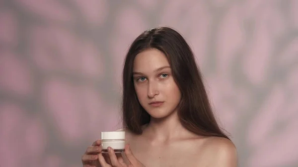 化粧品を保有する若手モデルの肖像画 長い髪と自然なメイクでカメラを見ている女の子 ピンクのバックグラウンドで孤立 スキンケア広告コンセプト — ストック写真