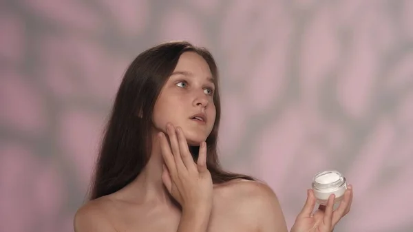 化粧品を保有する若手モデルの肖像画 長い髪とヌードの化粧をした可愛い女の子 ピンクのバックグラウンドで孤立 スキンケア広告コンセプト — ストック写真