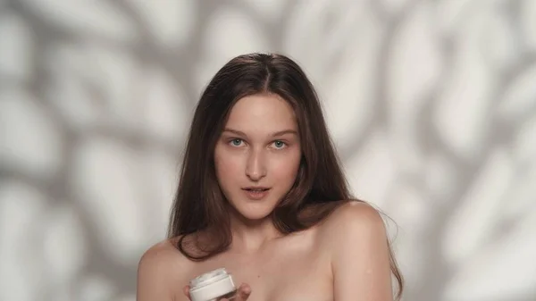 一个持有化妆品的年轻模特的画像 头发长 妆容柔和的女孩看着镜头笑了 被白色背景隔离 Skincare广告概念 — 图库照片