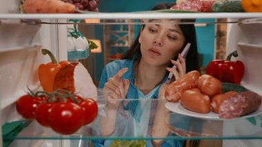 Buzdolabında kulağına telefon dayanmış Asyalı bir kadın. Bir kadın buzdolabındaki yiyecekleri inceliyor ve sipariş veriyor. Satın almak, yemek sipariş etmek. Buzdolabının içinden görüntüle