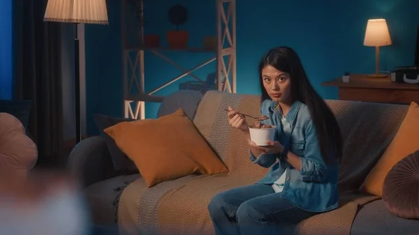 一位亚洲妇女正坐在客厅的沙发上 用勺子从桶里吃着冰淇淋 那女人下巴上沾满了冰淇淋 惊讶地直视前方 — 图库照片