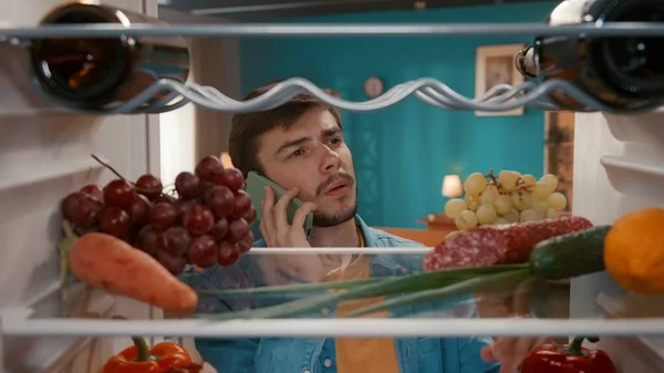 stock image A man with a phone to his ear at the open refrigerator. A man inspects the food in the refrigerator, and makes an order for delivery. Purchase, order foods. View from inside the refrigerator