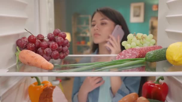 Eine Asiatin Mit Einem Telefon Ohr Öffnet Die Kühlschranktür Eine — Stockvideo