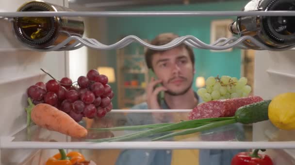 Mężczyzna Telefonem Przy Uchu Otwiera Drzwi Lodówki Mężczyzna Sprawdza Jedzenie — Wideo stockowe