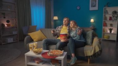 Genç romantik bir çift film izliyor ve patlamış mısır yiyor. Gülümseyen adam ve kadın oturma odasında rahat bir kanepede sarılıp dinleniyorlar, komedi filminin tadını çıkarıyorlar. HDR BT2020 HLG Malzemesi