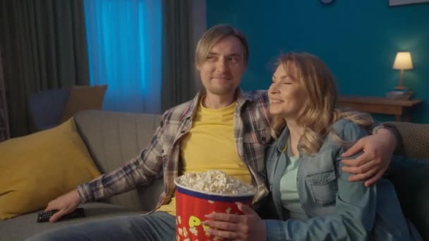 若いカップルが映画を見てポップコーンを食べている 笑顔の男女がリビングで快適なソファーで抱擁し リラックスしてコメディ映画を楽しんでいます 愛する夫婦の肖像画 — ストック動画