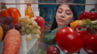 Asyalı bir kadın sağlıklı ürünlerle dolu bir buzdolabını inceliyor: sebze, meyve, böğürtlen. Tatmin olmuş bir kadın, açık buzdolabının yanında, tatmin olmuş bir şekilde başını sallar. İçeriden görüntüle