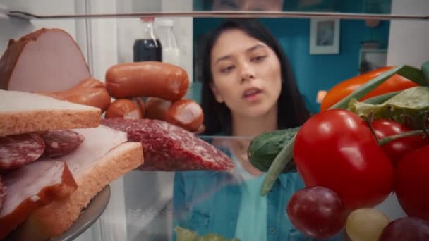 一位亚洲妇女站在一个开着的冰箱旁 冰箱里有健康和不健康的产品 女人耸了耸肩 拿不定主意吃什么 从冰箱里面看 — 图库视频影像