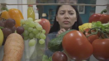 Düşünceli, kafası karışmış genç bir Asyalı kadın sağlıklı yiyeceklerle dolu açık bir buzdolabının yanında duruyor. Kadın çenesini tutuyor ve başını sallıyor. Atıştıracak bir şey bulamıyor. İçeriden görüntüle