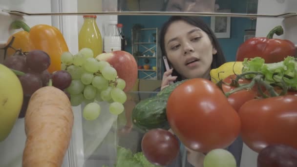 Азиатка Стоит Открытого Холодильника Телефоном Уха Женщина Осматривает Еду Холодильнике — стоковое видео