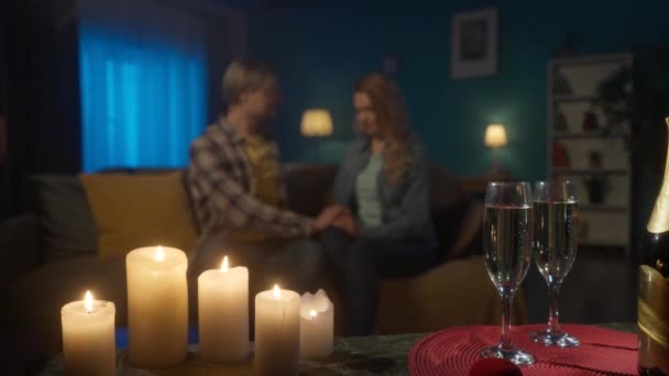 シャンパンのグラスと背景のぼやけた愛情のあるカップル リビングルームのソファに座っている間 愛の男と女がキスをしています シャンパンとロマンチックなキャンドルライトの夜 — ストック動画
