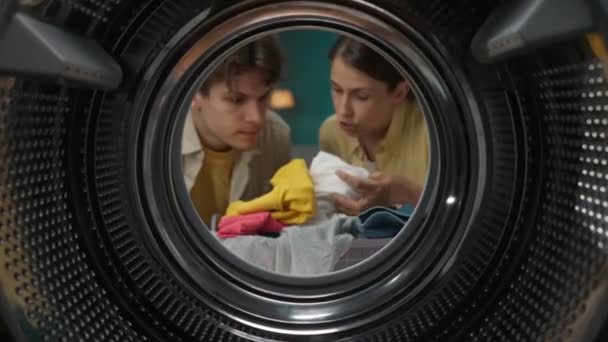 年轻夫妇在洗衣服 女人向她的男人解释和展示如何洗衣服和使用洗衣机 夫妻一起做家务活很开心 从空洗衣机的内部查看 — 图库视频影像