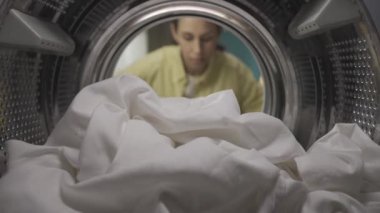Çamaşır sepetli genç bir kadın çamaşır makinesinin kapısını açıyor ve büyük beyaz çarşafı çıkarıyor. Bir şişe yumuşatıcı tutuyor, gülümsüyor ve göz kırpıyor. Pov çamaşır makinesinin içinde. HDR BT2020 HLG