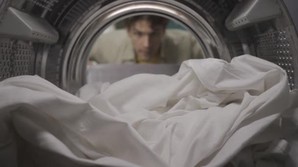 拿着洗衣篮的年轻人打开洗衣机的门 拿出大的白床单 他拿着一瓶织物柔软剂 笑了笑 竖起大拇指 洗衣机里的Povhdr — 图库视频影像