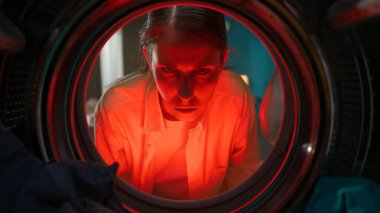 Çamaşır makinesinin içinde kıyafetlerle dolu kızgın görünen, gündelik giysiler içindeki genç bir kadının portresi. Kırmızı ışık alarmı. Çamaşır makinesinin içinden bak.