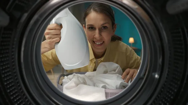 近距离拍摄的成年女性穿着休闲装 洗衣篮捧着一瓶柔软剂 面带微笑 从洗衣机内观看 — 图库照片