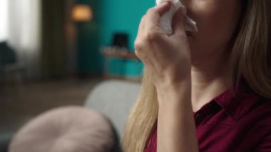 Sarışın bir kadının kanepede oturup burnunu kağıt mendille silerken yakın plan çekimleri. Burnuna burun spreyi sıkarak. Rhinitis tedavisi. Ev hekimliği konsepti. Sağlık hizmetleri reklamı.