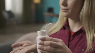 Sarışın bir kadının kanepede oturup, ilaç şişesinden bir hap alıp bir bardak suyla birlikte içtiği yakın çekim. Hastalık tedavisi. Ev hekimliği konsepti. Sağlık hizmeti reklamı. HDR