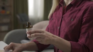 Kanepede oturan sarışın bir kadının yakın plan bir ilaç şişesini alıp plastik bir kapağa bir doz öksürük şurubu dökerken görüntüsü. Grip tedavisi. Ev hekimliği konsepti. Yavaş çekim HDR BT2020 HLG