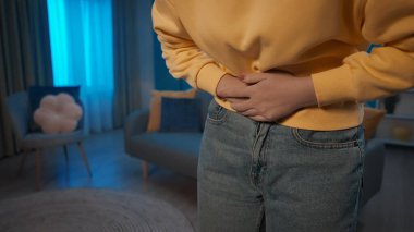 Oturma odasında dikilen bir kadın acı çekiyor. Karnını yakından tutan bir kadının elleri. Bir kadın karın ağrısı, regl ağrısı, hazımsızlık ve ishalden muzdarip. Ev hekimi konsepti