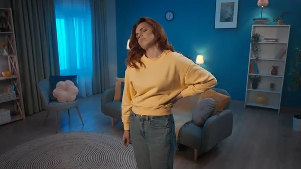 Redhaired woman suffers from back pain while standing in the living room. The woman arches, holding her back. A woman suffers from pain in her back, lower back, pinched nerve. Home medicine concept