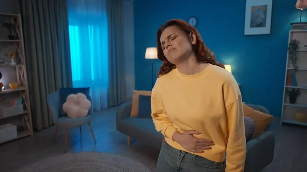 Bir kadın elini karnının üstünde tutuyor, acıdan kıvranıyor, oturma odasında duruyor. Bir kadın karın ağrısı, regl ağrısı, hazımsızlık ve ishalden muzdarip. Ev hekimi konsepti