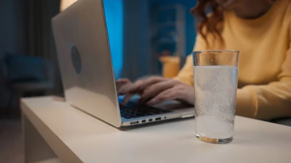 An effervescent tablet dissolves in a glass of water on a table close up. In the background, a womans hands are typing on a laptop. Cure for a cold, pain or hangover. Home medicine concept