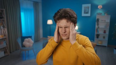 Mutsuz üzgün adam oturma odasında şakaklarına masaj yapıyor, yüzünü buruşturuyor, şiddetli baş ağrısı çekiyor, kapat. Gerilim, migren, yorgunluk yüzünden insan kendini kötü hisseder. Ev hekimi konsepti