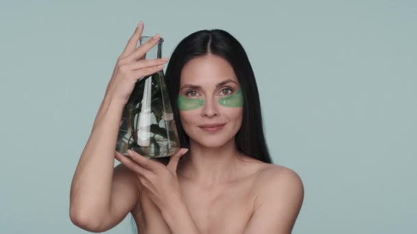 一个眼皮底下有绿色水凝胶斑斑的女人拿着一个装有植物的瓶子 穿着蓝色背景衣服在演播室显赫的女人 以天然植物成分为原料的化妆品产品 — 图库视频影像